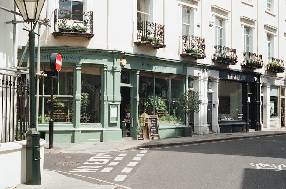 An exterior view of a charming flower shop with a pale green facade named 'Kensington Flowers,' situated on a corner of a street near Kentish Town Road NW5. The shop features large display windows fil