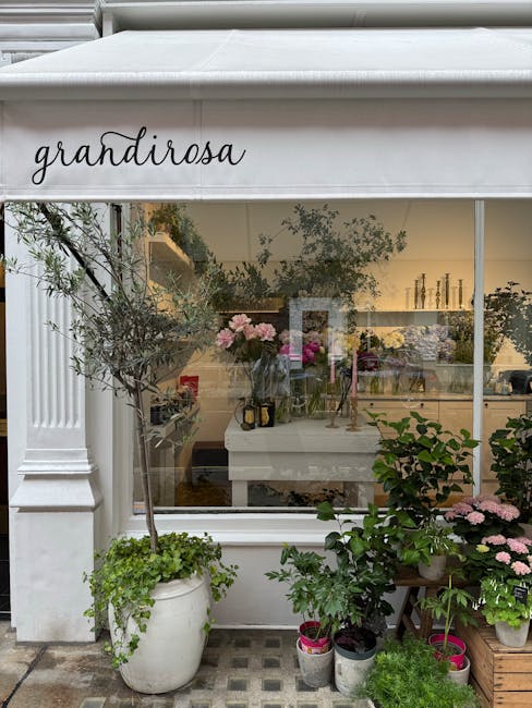 A floral shop storefront named 'grandirosa' featuring a large glass window with a white frame. Outside, there are potted plants, including a small tree with slender branches and leafy foliage in a whi