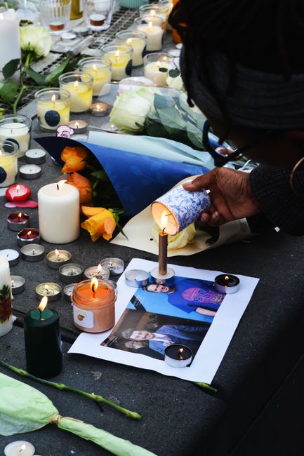 A person with dark skin and black hair wearing glasses is lighting a small candle placed among a floral tribute on a black surface. The tribute includes white roses, orange flowers, and greenery, wrap