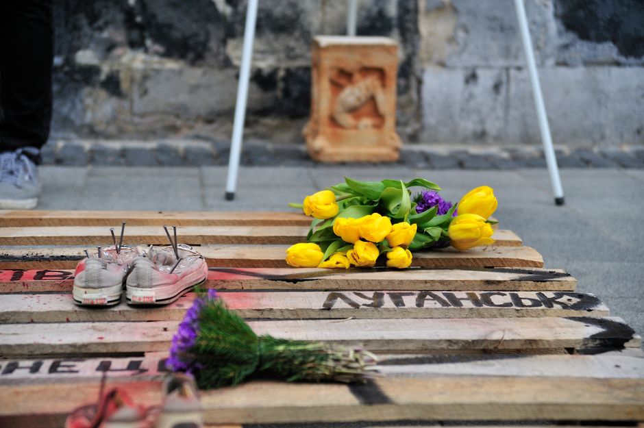 A floral tribute consisting of a bouquet of bright yellow tulips and purple flowers resting on a wooden pallet with painted black and red markings. Nearby, a pair of white sneakers with laces and pins