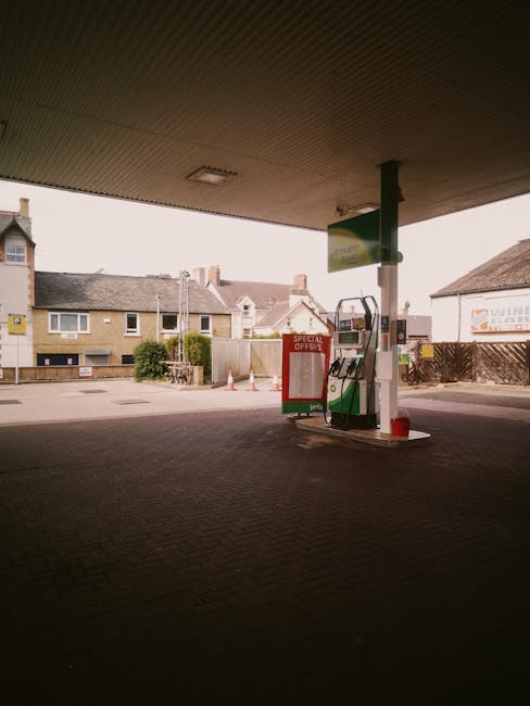 A view from underneath a petrol station canopy shows a green and white fuel pump with a small red cone placed nearby. In the background, residential houses with brick and light-colored walls are visib