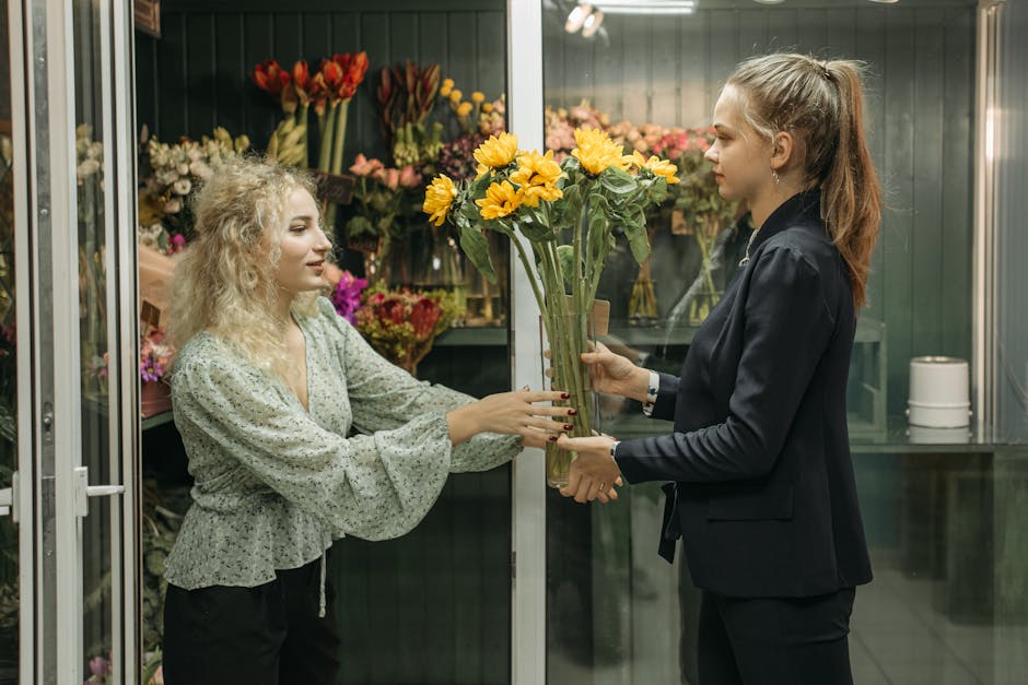 A woman with curly blonde hair wearing a light green floral blouse is handing a bouquet of bright yellow daisies with green stems and leaves to a flower shop assistant with straight brown hair tied ba