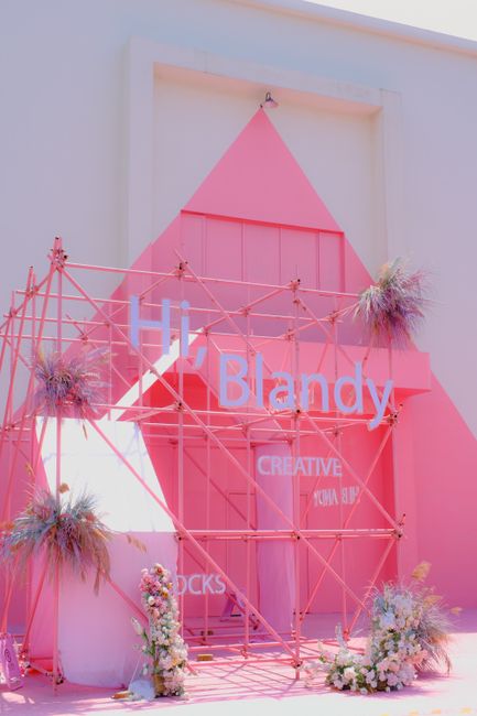 A decorative pink geometric backdrop featuring an arrangement of fresh flowers at the base, including white and pink blossoms with green foliage, complemented by small, spiky, purple-hued plants. The 