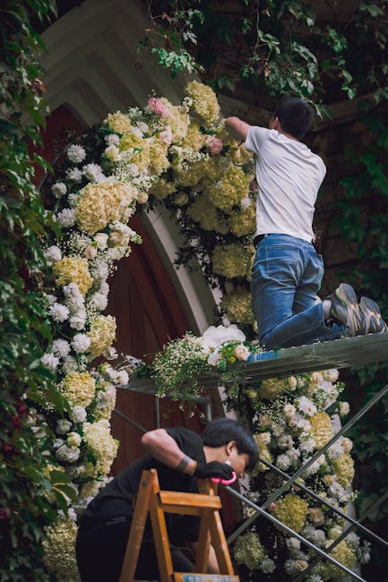 A person in a white shirt and blue jeans kneels on a scaffold, reaching up to arrange a floral display on the exterior wall of a building. The floral installation features a large, semi-circular arch 