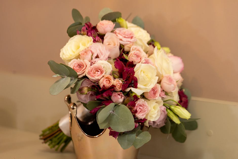 A bouquet arrangement featuring a mix of fresh, pastel pink and white roses, complemented by deep burgundy flowers and silvery-green eucalyptus leaves. The flowers are tightly clustered and appear rec
