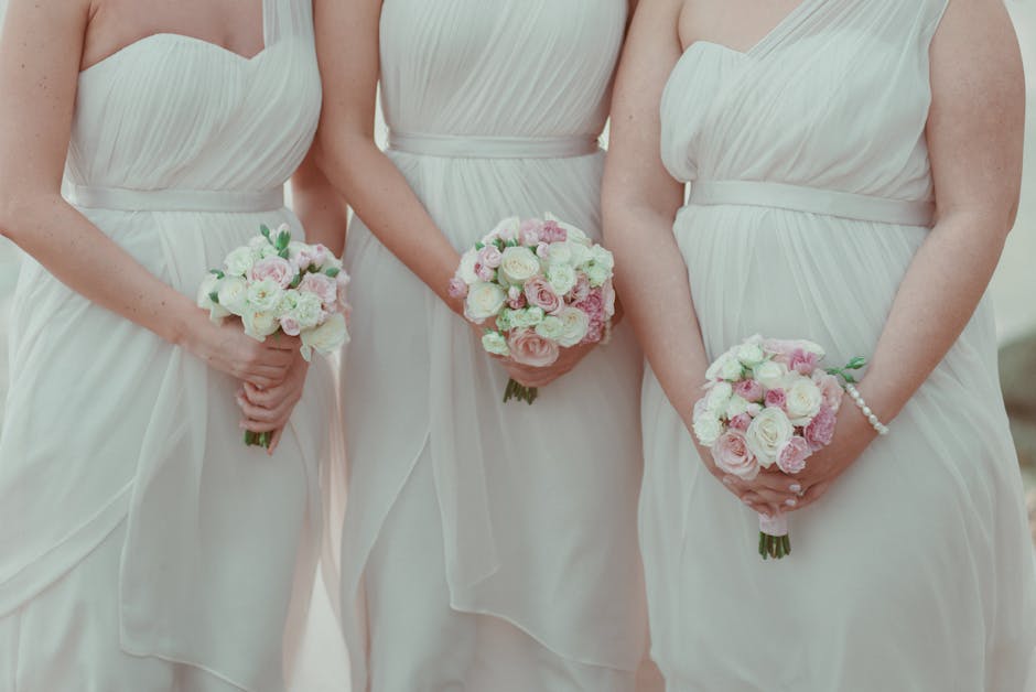 Close-up of three women standing side by side, each holding a bridal bouquet arranged with white and light pink roses, peonies, and greenery. They are dressed in elegant, white, sleeveless wedding dre