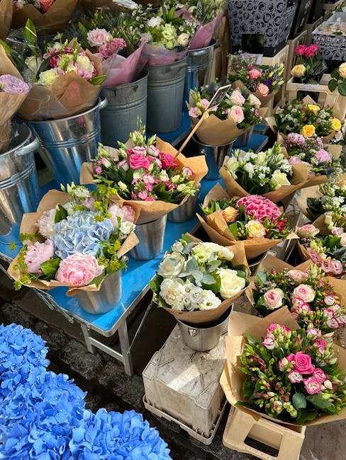 A vibrant display of fresh flower bouquets arranged in metal buckets and paper wrappers on a market stall, featuring a variety of pink, white, and blue blooms including roses, hydrangeas, and carnatio