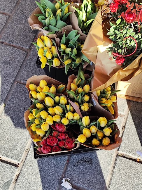 Three bouquets of fresh flowers arranged in brown paper wrapping and placed on a paved outdoor surface. The first bouquet features vibrant yellow tulips with closed buds and green leaves. The second b