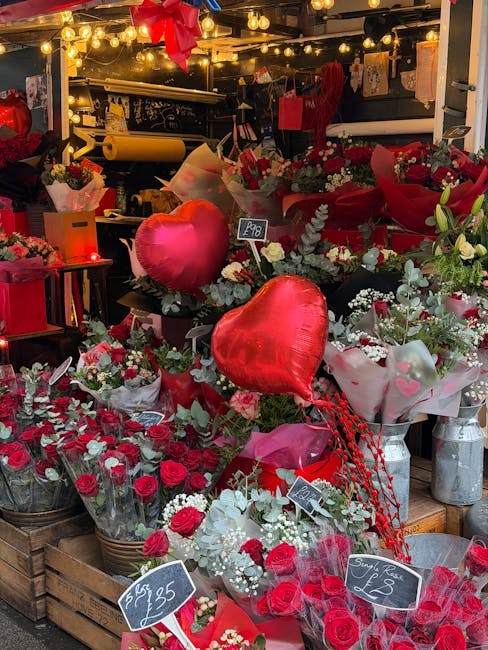 A display of fresh floral arrangements and bouquets in a florist shop, featuring red roses, white flowers, and greenery, arranged in baskets and wrapping paper. The display includes several heart-shap
